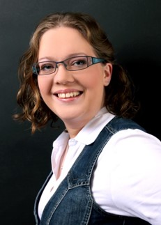 Portraitfoto der Social-Media-Beratern Sarah Schückel, einer Frau mit offenen Haaren in weißem Shirt und Jeansweste vor dunklem Hintergrund.